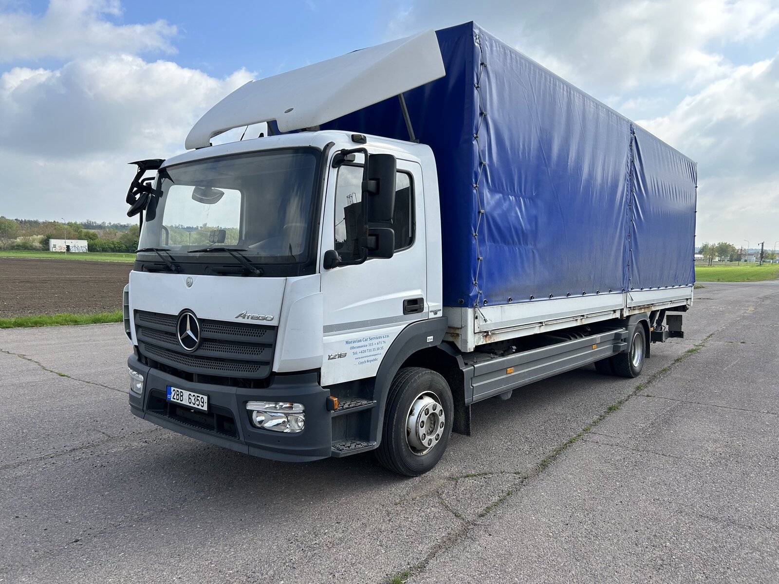 Bílý nákladní vůz Mercedes-Benz Atego s modrou plachtovou nástavbou